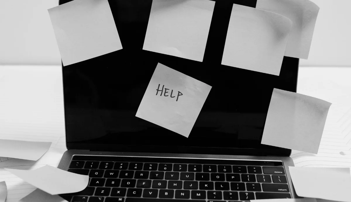 A laptop screen obscured by loose handwritten notes, one of them reading HELP